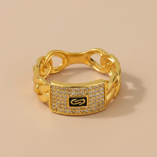 A gold chain-link ring with a rectangular pavé-set centerpiece featuring a black background and a gold infinity symbol, accented with small clear stones.