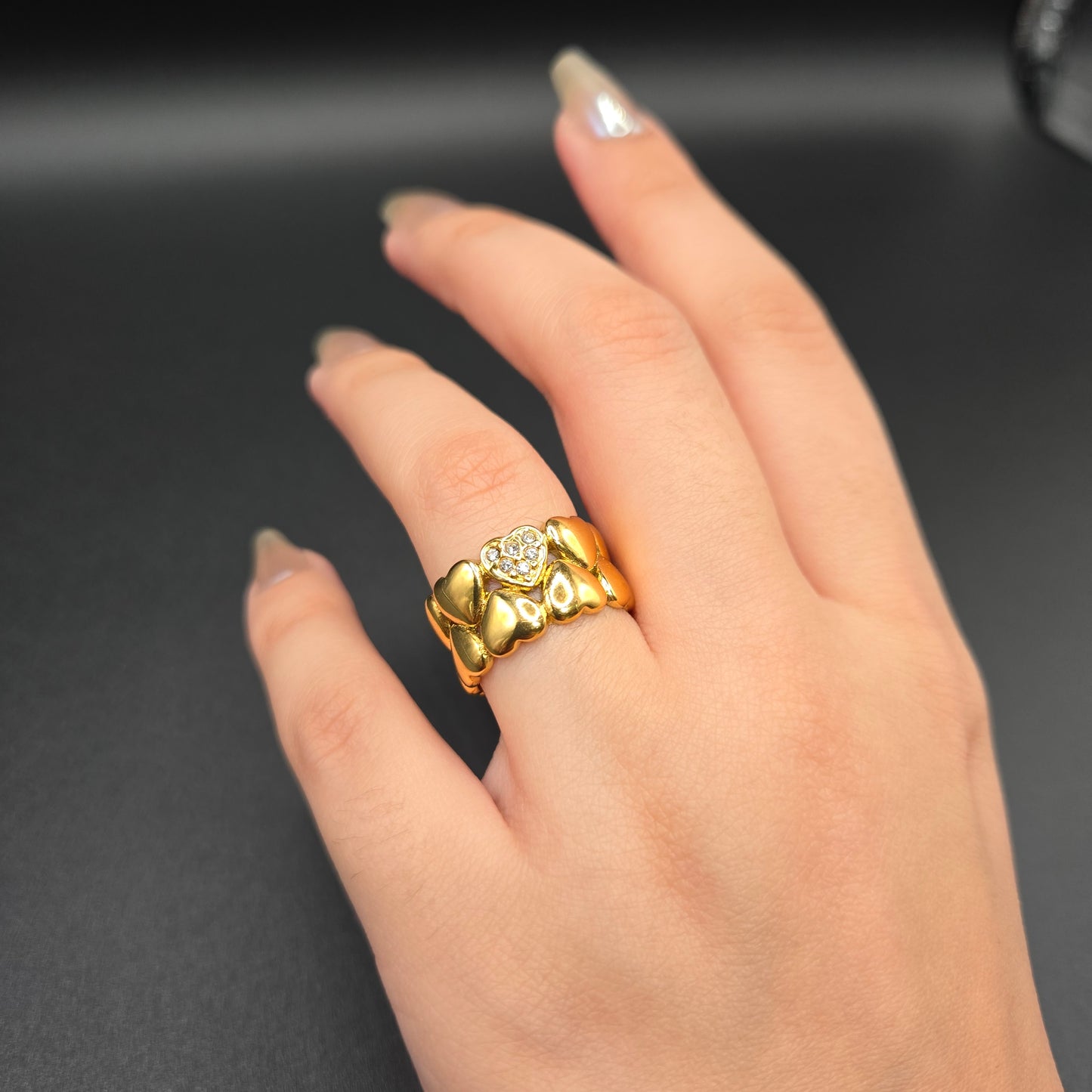 A person's hand showcasing a gold-colored ring with a heart pattern and a shiny stone on the finger.