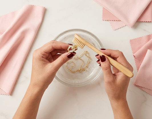 Woman's hands gently cleaning a gold-plated knot ring with a soft brush and soapy water to prevent tarnishing.