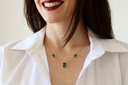 a woman wearing a delicate gold necklace with three small rectangular green gemstones, styled with a white collared shirt.