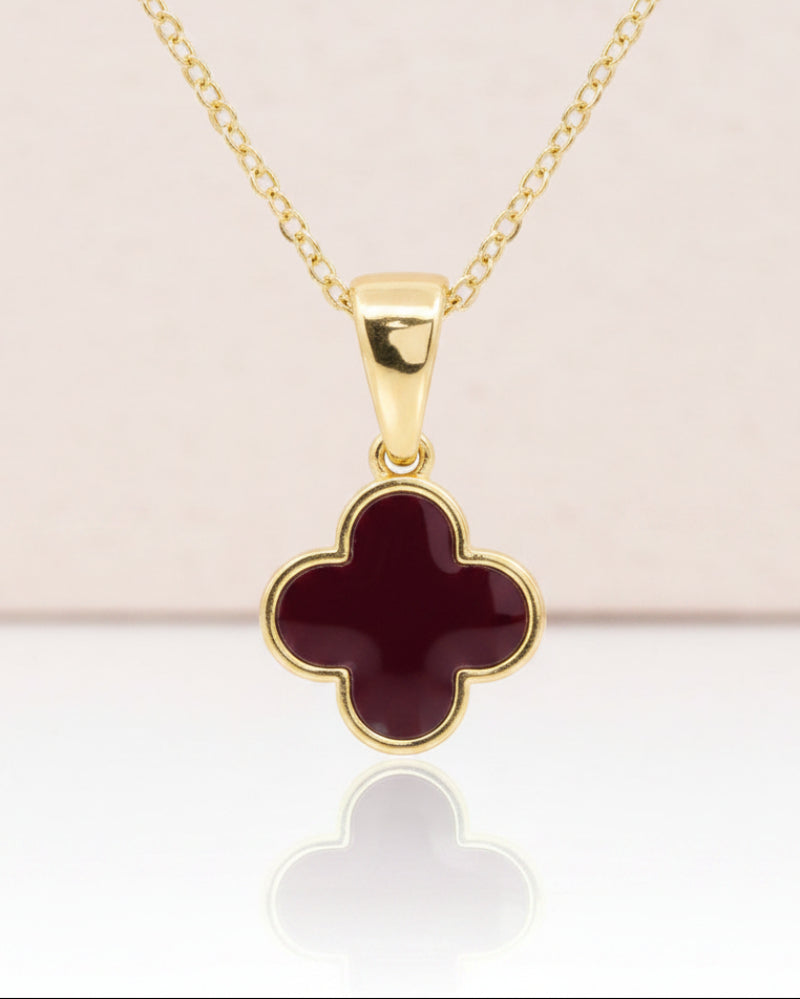 A gold chain necklace with a four-leaf clover pendant featuring a burgundy inlay and gold border, displayed on a clean white background with a soft reflection below.