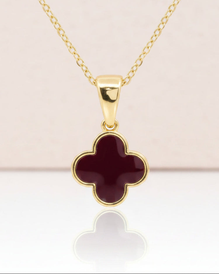 A gold chain necklace with a four-leaf clover pendant featuring a burgundy inlay and gold border, displayed on a clean white background with a soft reflection below.