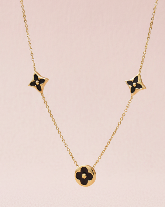 A gold-tone necklace featuring four small monogram flower charms with black enamel details and a larger clover pendant at the center, displayed against a soft pink background.