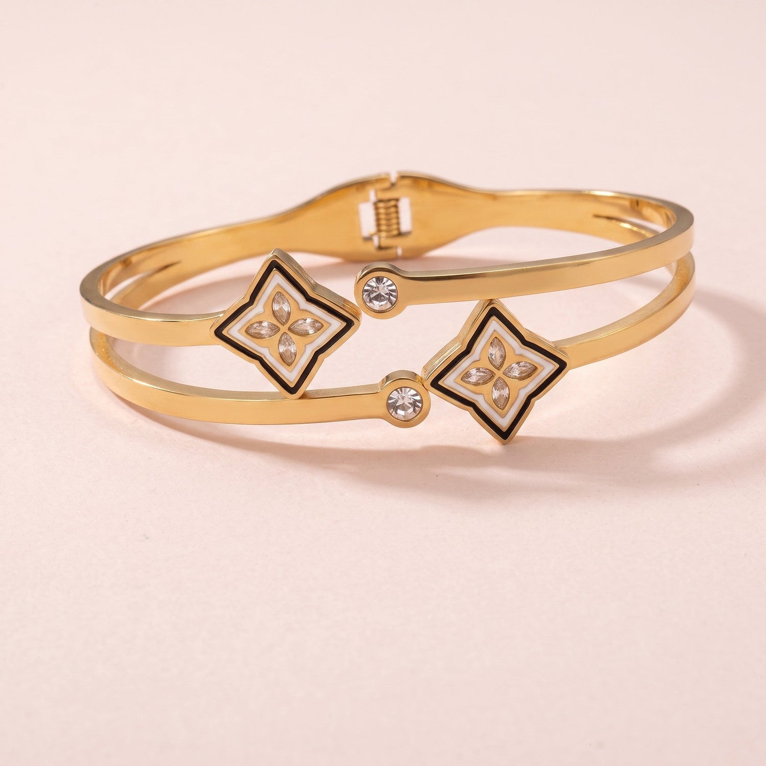 A gold-tone necklace featuring four small monogram flower charms with black enamel details and a larger clover pendant at the center, displayed against a soft pink background.