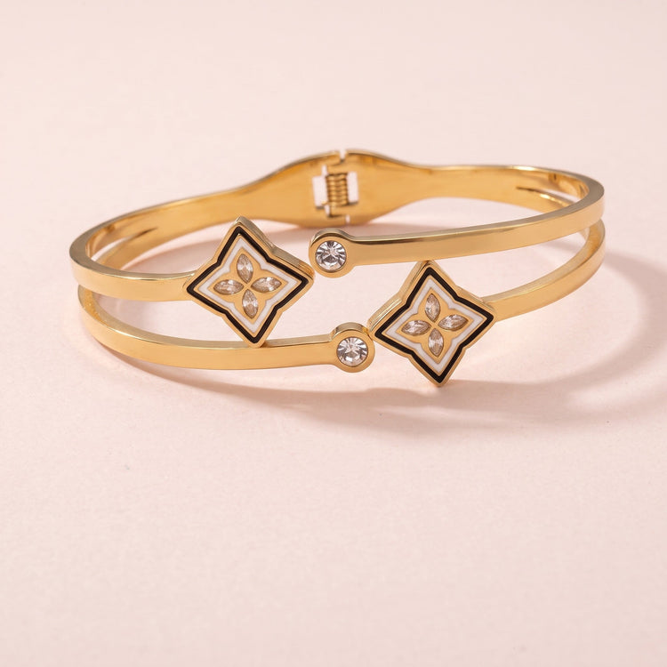A gold-tone necklace featuring four small monogram flower charms with black enamel details and a larger clover pendant at the center, displayed against a soft pink background.