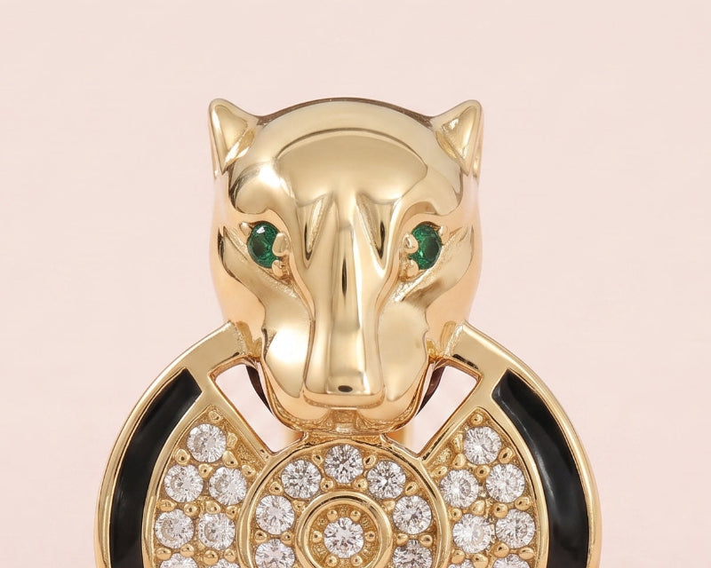 A yellow gold Panther ring, featuring a panther head with green cubic zirconia eyes, a black enamel collar, and a circular pavé motif on a soft pink background.