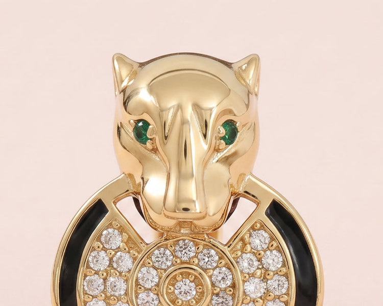 A yellow gold Panther ring, featuring a panther head with green cubic zirconia eyes, a black enamel collar, and a circular pavé motif on a soft pink background.