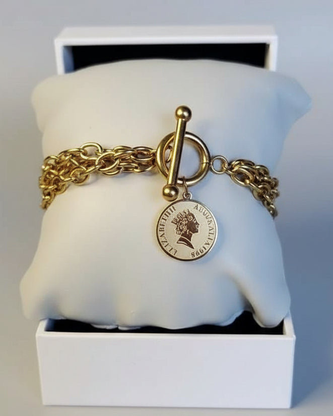 A gold-tone toggle clasp bracelet with a round Queen Elizabeth II coin charm, displayed on a white padded jewelry pillow inside an open white box.