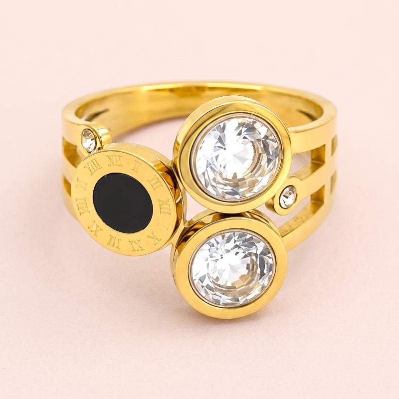 A gold-plated ring featuring three circular bezel settings: two clear faceted gemstones and one round black gemstone, with small accent crystals and Roman numeral details on the band, set against a soft pink background.