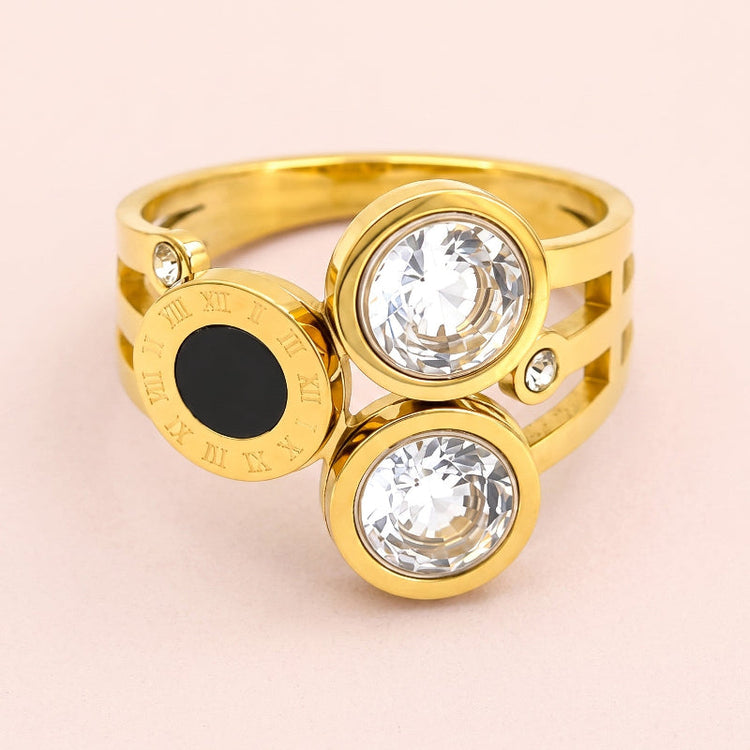 A gold-plated ring featuring three circular bezel settings: two clear faceted gemstones and one round black gemstone, with small accent crystals and Roman numeral details on the band, set against a soft pink background.