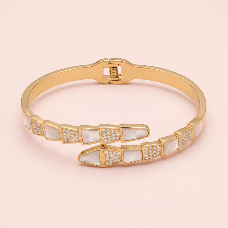 A gold bangle bracelet featuring serpent design mother-of-pearl inlay segments, some embellished with small clear pavé stones, shown on a soft pink background.