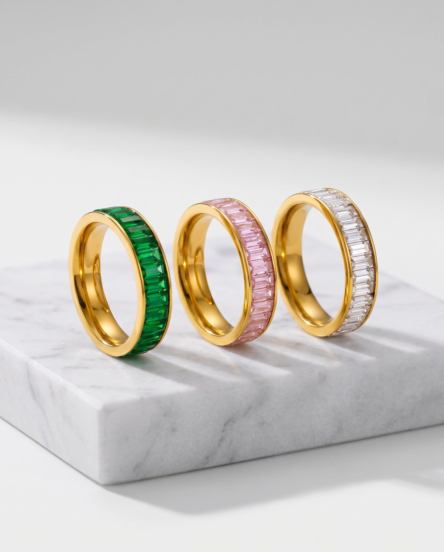 Three gold rings with textured, ridged bands are displayed on a warm beige surface. The left ring has a vibrant green inlay, the middle ring has a soft pink inlay, and the right ring has a white inlay.