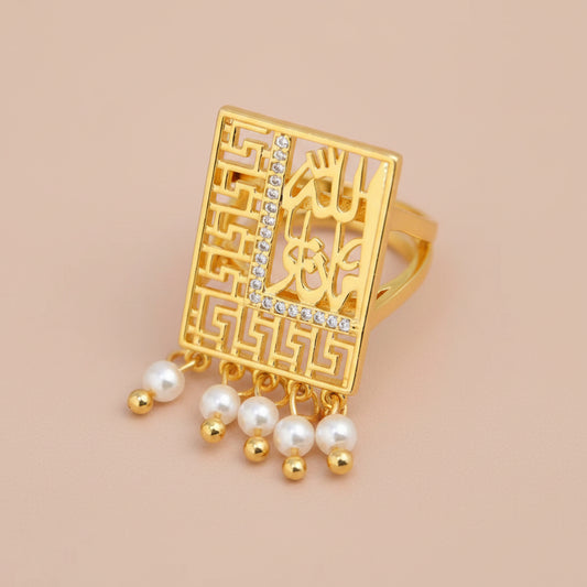 A gold-plated rectangular ring featuring intricate filigree cutouts, geometric border patterns, and dangling pearl beads with small gold beads, set against a soft white satin background.
