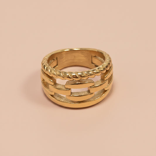 A gold chain-link ring with a twisted rope accent band, photographed on a clean pink background.
