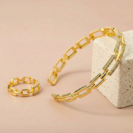A gold-toned chain bracelet and matching ring with rectangular links and small clear stones, displayed on a flat pink surface with a cream rock prop.