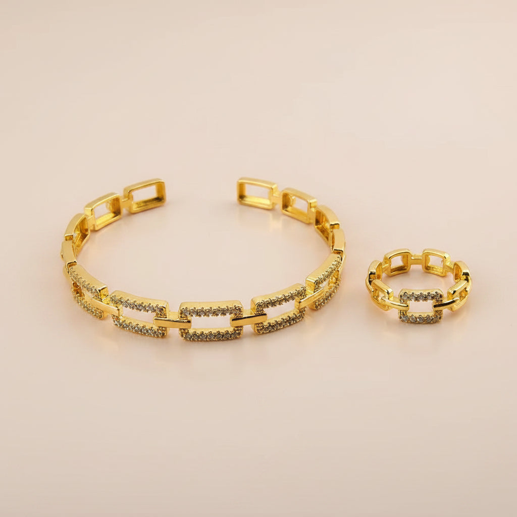 A gold-tone link bracelet and matching ring set, both decorated with rows of small clear stones, displayed on a flat pink surface