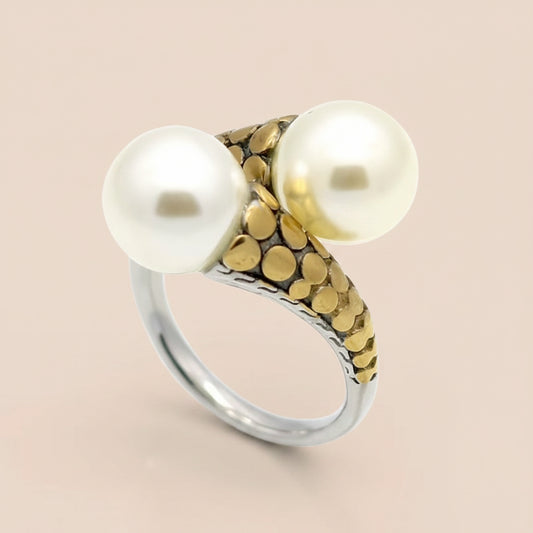 A unique ring featuring two large white pearls set in a textured gold V-shaped band with a polished silver-toned lower band, shown on a pink background.