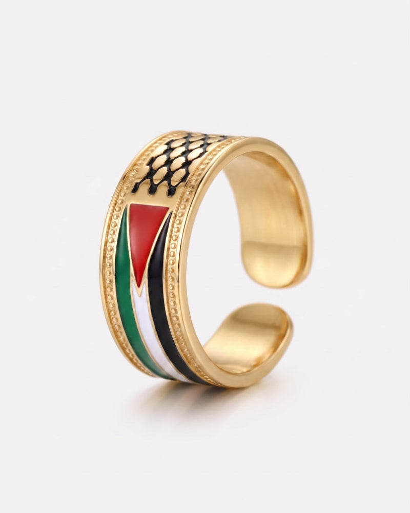 A gold-colored open cuff ring featuring an inlaid design of the Palestinian flag (red, white, green, black) and a black fish-scale pattern on top, set against a light gray background.
