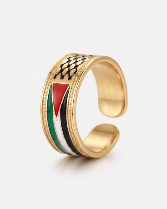 A gold-colored open cuff ring featuring an inlaid design of the Palestinian flag (red, white, green, black) and a black fish-scale pattern on top, set against a light gray background.