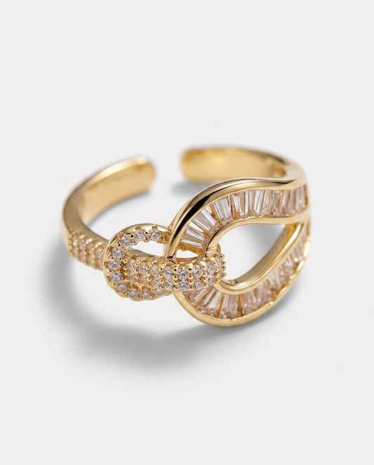 A gold-toned adjustable ring featuring a knotted design embellished with clear baguette and round crystals, set against a light gray background.