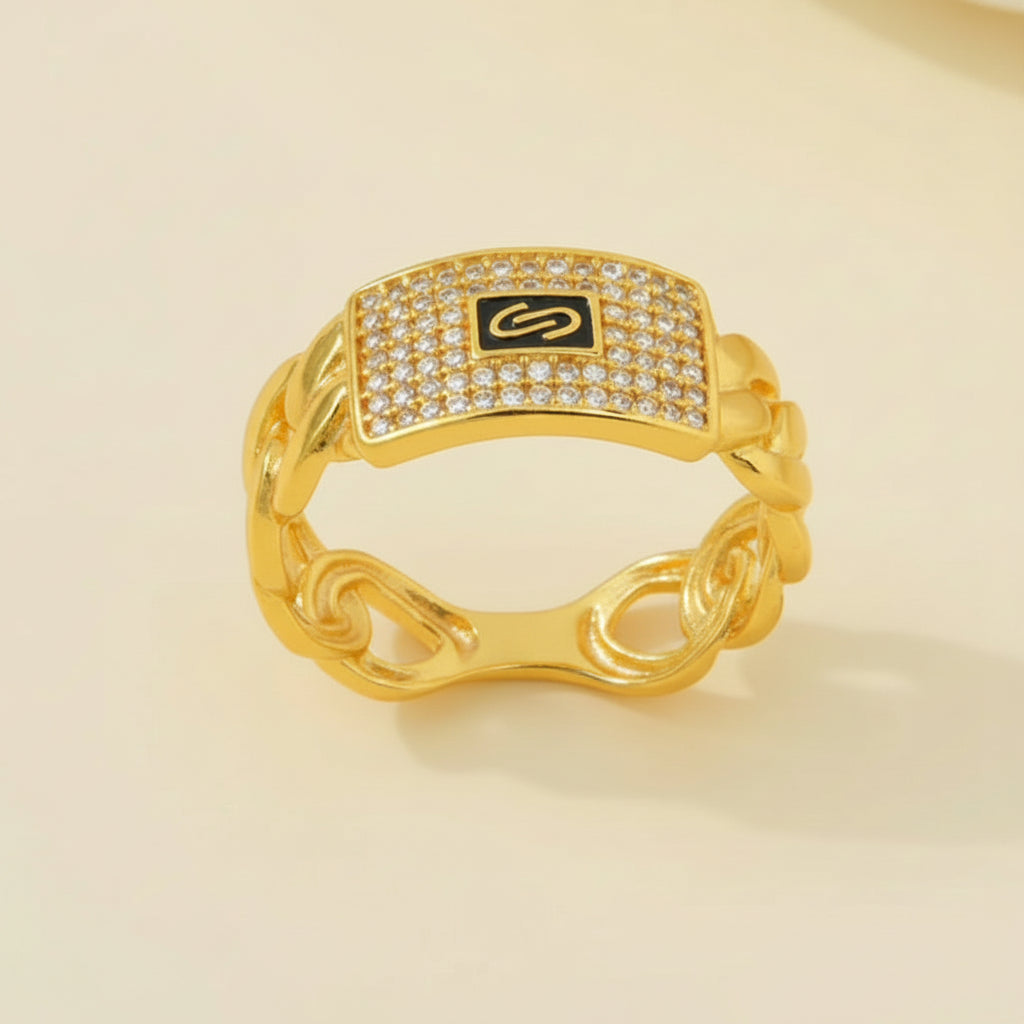 A gold chain-link ring with a rectangular face paved with clear stones and a black center featuring a gold monogram logo, set against a light beige background.