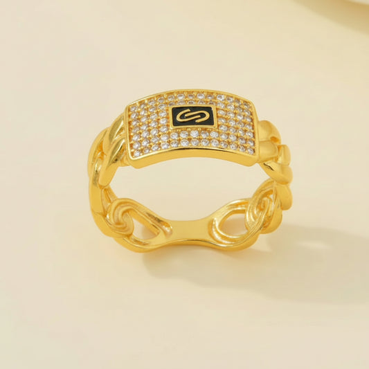 A gold chain-link ring with a rectangular face paved with clear stones and a black center featuring a gold monogram logo, set against a light beige background.