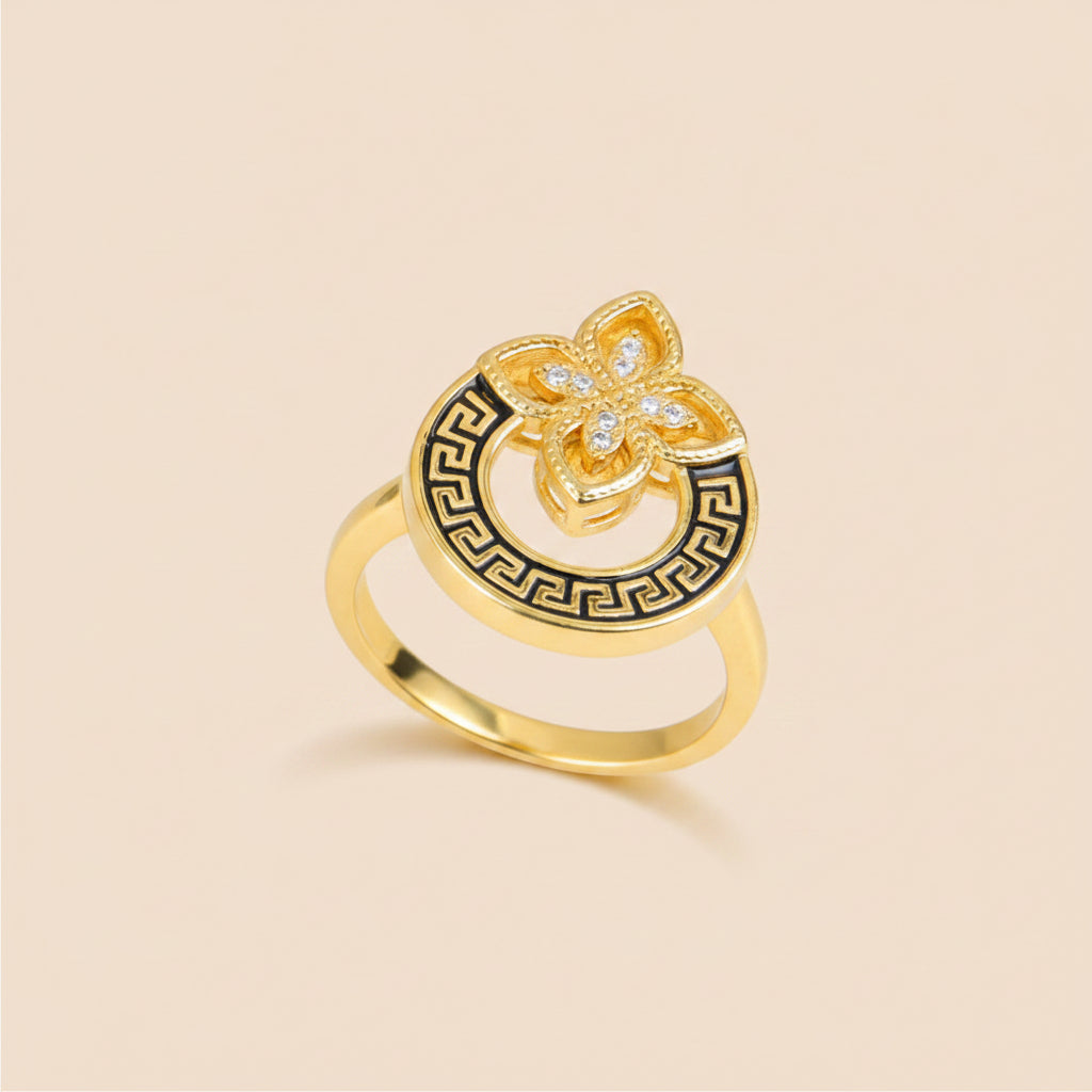 A gold ring with a circular Greek key pattern and a central flower motif set with small clear stones, displayed on a beige curved surface.