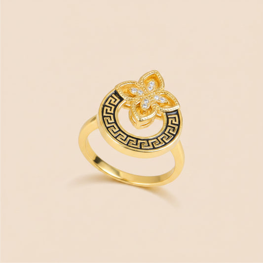 A gold ring with a circular Greek key pattern and a central flower motif set with small clear stones, displayed on a beige curved surface.