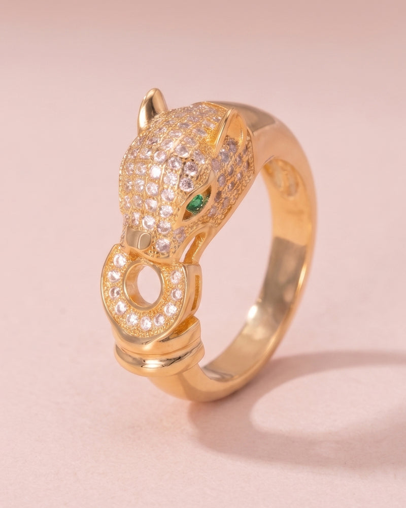 A gold ring shaped like a panther head, adorned with pavé-set clear stones and a small green eye accent, photographed on a soft pink background.