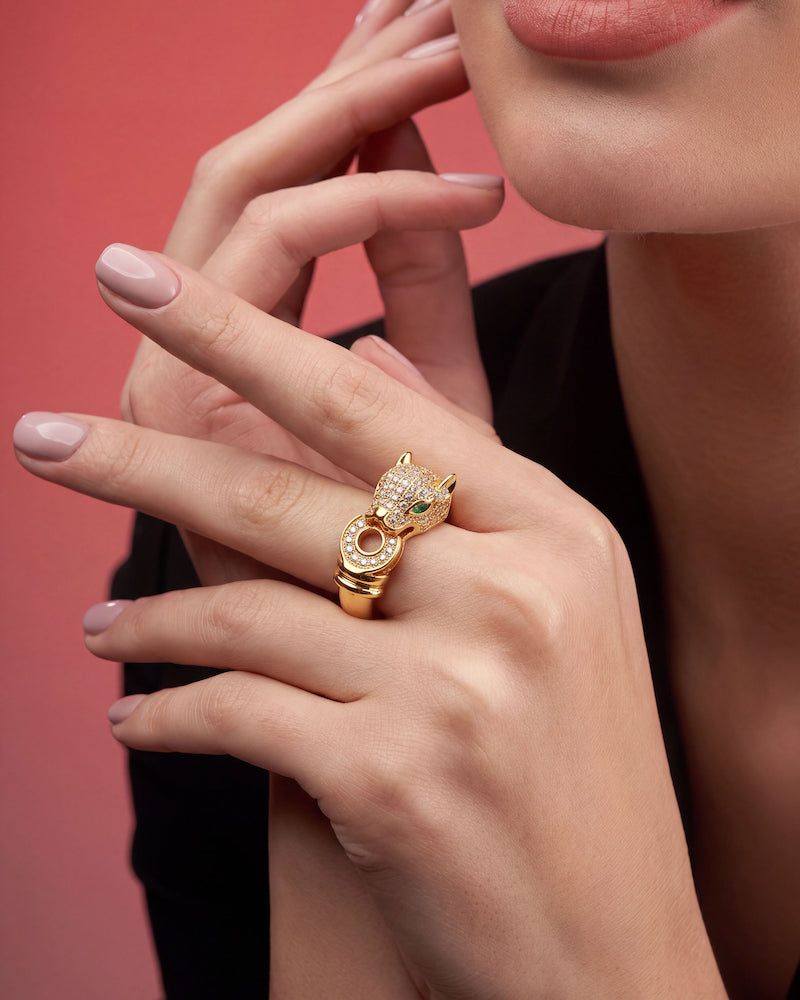 A close-up lifestyle shot of a model wearing a gold ring shaped like a panther's head, adorned with small clear crystals and a tiny green gemstone, set against a solid pink background.