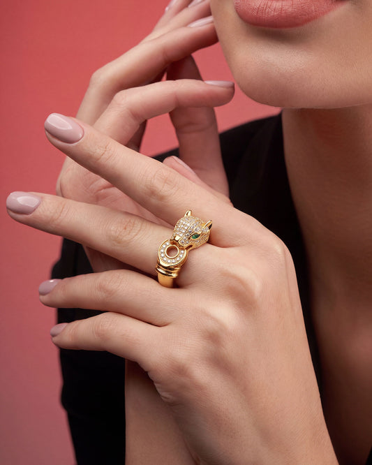 A close-up lifestyle shot of a model wearing a gold ring shaped like a panther's head, adorned with small clear crystals and a tiny green gemstone, set against a solid pink background.