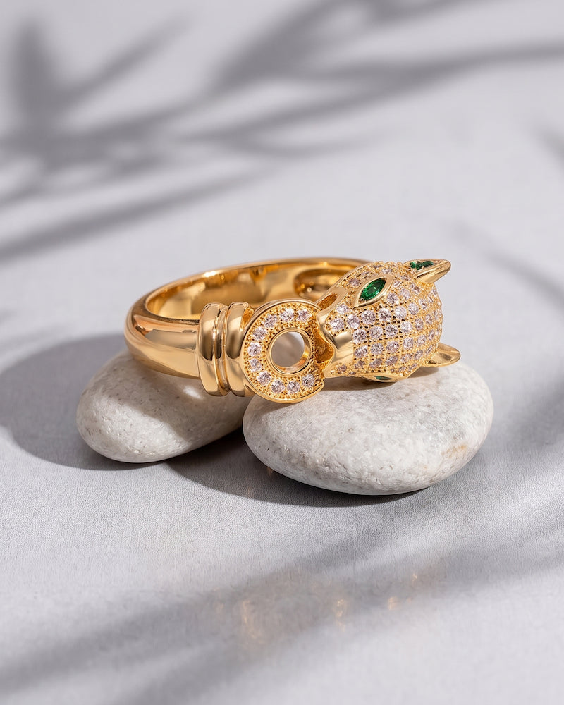 A gold ring shaped like a panther, decorated with pavé-set clear stones and small green gemstone eyes, displayed on two smooth white stones against a light gray fabric background with soft leaf shadows.