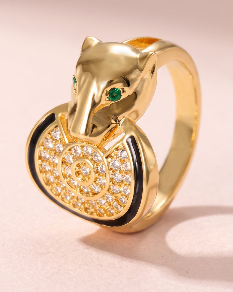 A gold ring shaped like a panther head holding a heart-shaped cluster of clear pavé stones with black enamel accents and small green stone eyes, shown on a soft pink background.