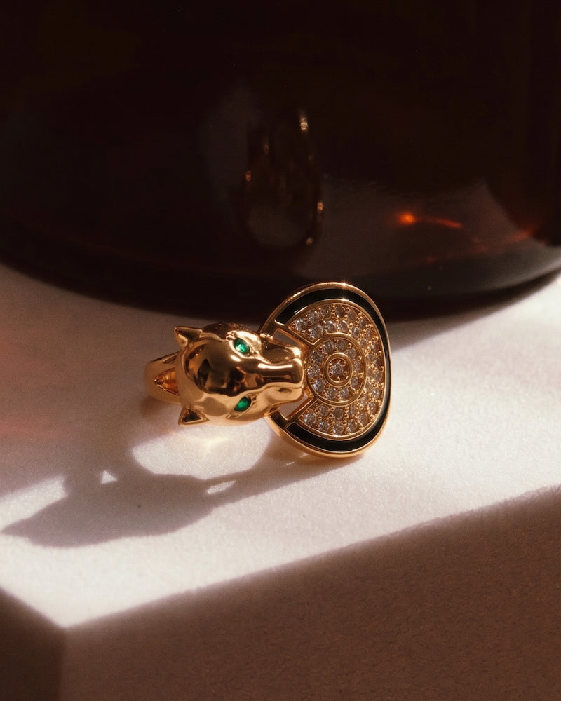 A gold ring shaped like a panther head with green eye accents and a circular pavé face, resting on a white surface in dramatic warm lighting.