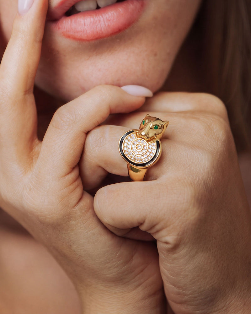 A close-up lifestyle shot of a model wearing a gold ring shaped like a panther head with green enamel eyes and a diamond-encrusted circular face.