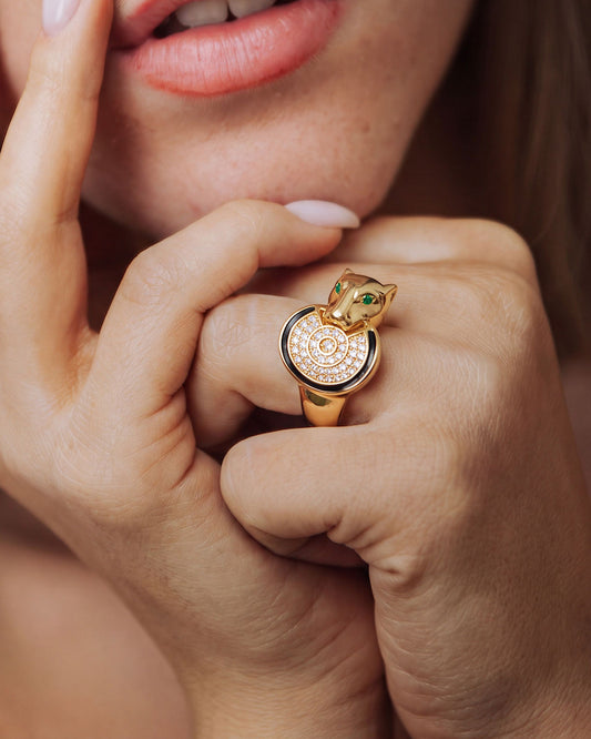 A close-up lifestyle shot of a model wearing a gold ring shaped like a panther head with green enamel eyes and a diamond-encrusted circular face.