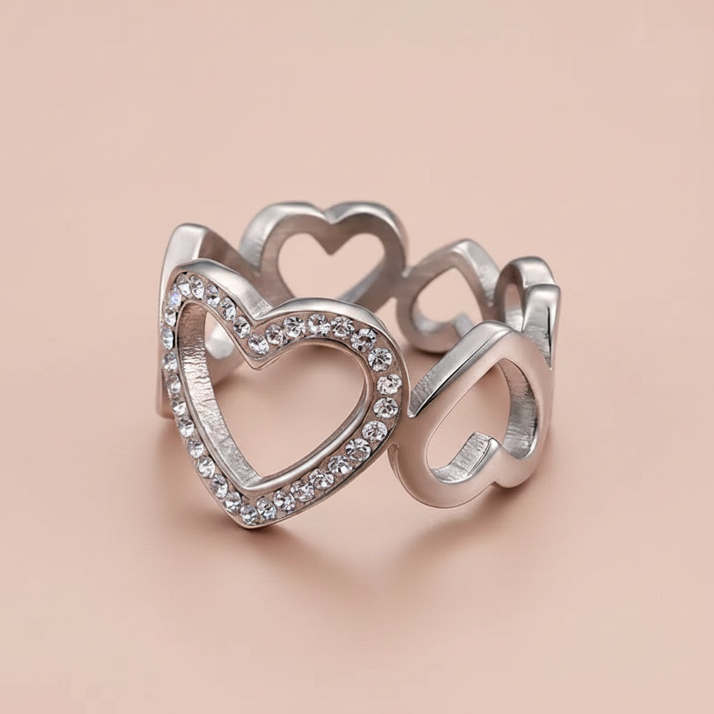 A silver-toned open heart ring with a front heart outlined in small clear stones, shown on a clean pink background.