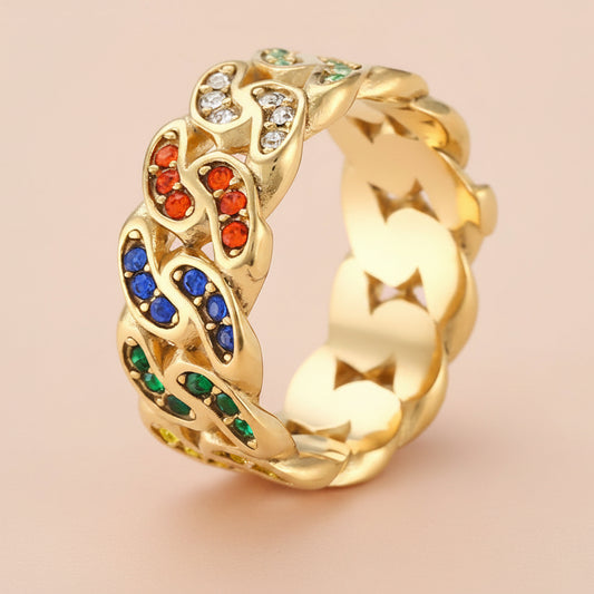 A gold-plated chain-link ring decorated with colorful rhinestones in red, blue, green, and white, set against a solid pale pink background.