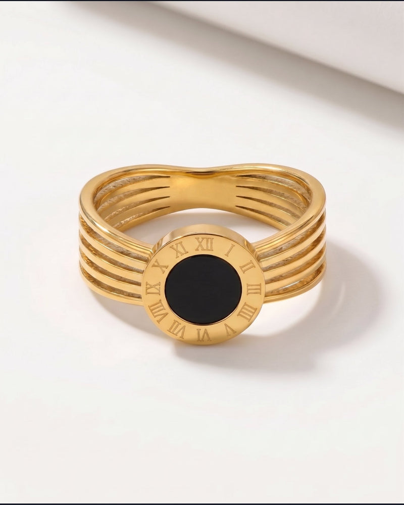 A gold-plated ring with a multi-strand band design, Roman numerals on the face, and a central black oval inlay, shown on a clean white background.