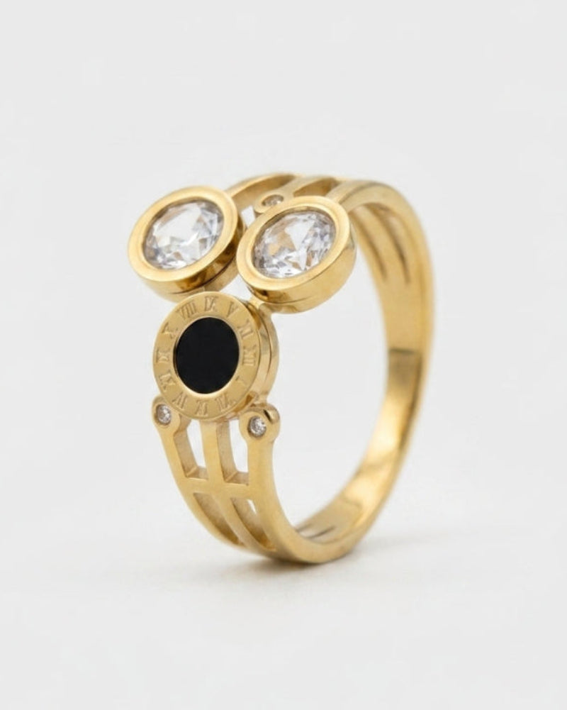A gold ring featuring two round bezel-set clear stones on top and a central oval black stone, photographed on a clean light gray background.