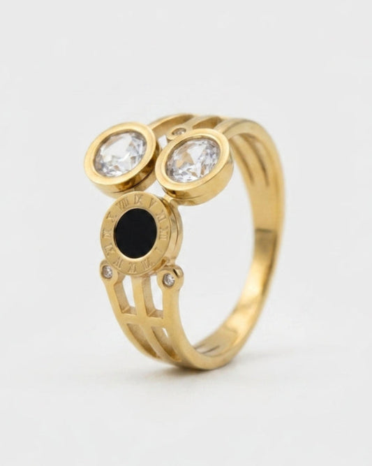 A gold ring featuring two round bezel-set clear stones on top and a central oval black stone, photographed on a clean light gray background.