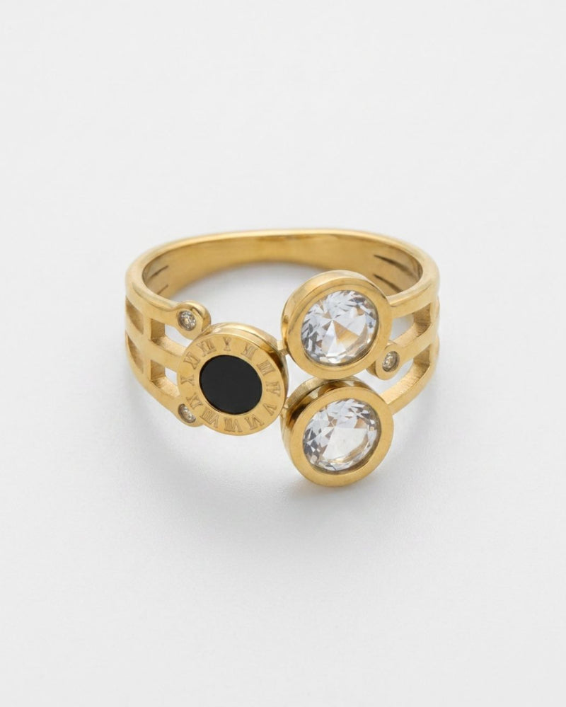 A gold ring with a geometric design, featuring two bezel-set clear round gemstones, a central round black gemstone, and small accent stones on a light gray background.
