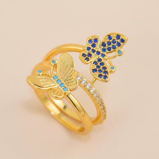 Two gold butterfly rings with blue and clear crystal accents, displayed on a soft beige surface with white flowers in the background.