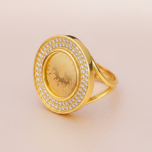A gold lira coin ring with a round face bordered by small clear stones, shown on a dark reflective surface. 