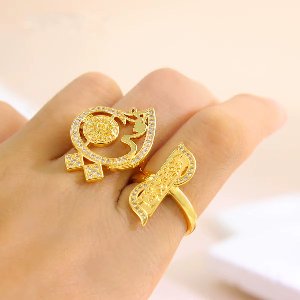 Two gold rings with clear crystal accents, one shaped like a stylized Om symbol and the other like a curved decorative piece, displayed on a person's hand against a soft beige background.