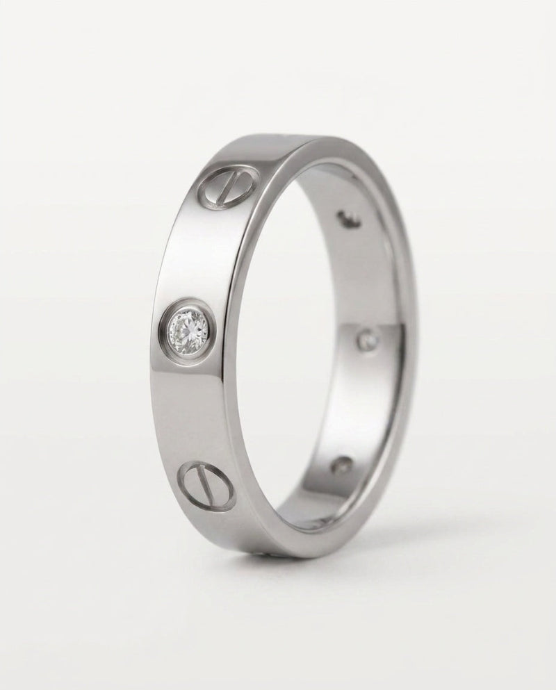 A silver-toned screw-ring band ring with three screw details and one clear gemstone, shown on a light gray background.