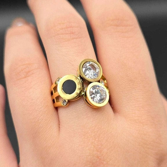 A gold ring with three circular settings worn on a finger. Two settings contain clear round gemstones, and one setting has a black oval inlay.